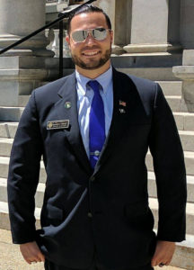 Brandon Phinney (NH rep) standing in front of legislative building in New Hampshire, wearing suit, tie, sunglasses, name badge, American flag lapel pin
