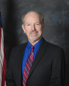Jeffrey Hewitt portrait standing in dark suit and red tie in front of American flag (color photo)