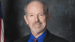 Jeffrey Hewitt portrait standing in dark suit and red tie in front of American flag (color photo)