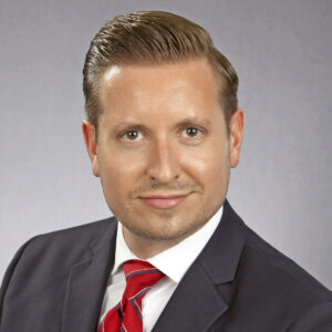 Drew G. Miller professional headshot, smiling, wearing dark grey suit, white shirt, red tie (color photo) 