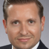 Alt text Drew G. Miller professional headshot, smiling, wearing dark grey suit, white shirt, red tie (color photo)