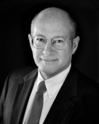Martin Cowen portrait, wearing dark suit and tie, eyeglasses, looking at camera, dark backdrop as in a photo studio (black and white photo)