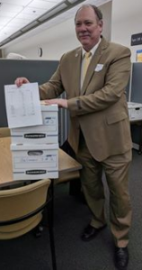 Bill Gelineau in tan suit standing in office, posing with his box of petitions, 2018 candidate for governor of Michigan (color photo)