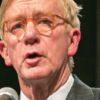 William Weld speaking at a microphone wearing grey suit and tie, eyeglasses, dark background (color photo)
