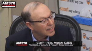 Murray Sabrin seated in radio studio, speaking on camera, microphone in front of him, wearing eyeglasses and dark sportcoat, turned to off-camera radio host at his left, text on screen identifying Sabrin as 'Guest -- Prof. Murray Sabrin' and 'Professor of Business & Finance, Ramapo College', also station ID of "AM970 THE ANSWER' (color image)
