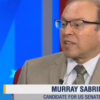 Murray Sabrin in tan suit & tie, wearing glasses, seat at TV studio desk, talking to hostess on his right (off camera), text on screen reads 'Murray Sabrin' and 'candidate for US Senate' and show title 'Power & Politics' (color photo)