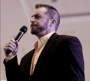 Adam Kokesh speaking at a mic wearing dark blazer, open-collar shirt (color photo)