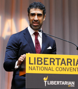Arvin Vohra in suit tie, speaking at lectern labeled 'Libertarian National Convention' and bearing the national Libertarian Party torch-eagle logo (color photo)