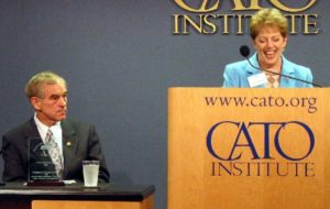 At left Ron Paul seated at desk on stage, at right Andrea Rich at lectern which reads 'www.cato.org' and 'CATO INSTITUTE' she is grinning as she presents him with a 2002 Thomas Szasz award (color photo)