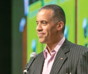 Larry Sharpe wearing grey pinstripe blazer, pale pink open-collared shirt, standing at microphone, smiling and looking toward audience, off-camera at viewer's left (color photo)