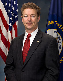 Sen. Rand Paul portrait, standing wearing grey suit red tie, smiling, in front of U.S. flag at left and Kentucky state flag at right (color photo)