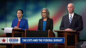 View of TV screen with three candidates from left Elinor Swanson, Kathleen Williams, Greg Gianforte all wearing suit jackets standing at black and brown lecterns, studio backdrop is projected, abstract large blurry US flag, label on TV screen is 'Tax cuts and the federal budget' (color image)