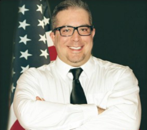 Richard Castaldo portrait, wearing white shirt, dark tie, eyeglasses, arms crossed, grinning, standing before an American flag on pole and dark backdrop (color photo)