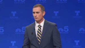 TV screen image of Kash Jackson speaking on stage at a mic wearing grey suit white shirt striped tie, blue backdrop featuring logos of Telemundo Chicago and NBC (peacock) Chicago Channel 5 (color image)
