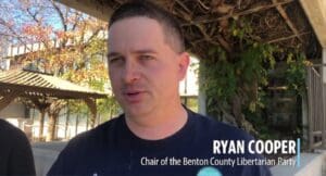 Video image of Ryan Cooper wearing navy blue T-shirt speaking to interviewer off-camera outdoors in front of building and trellis with white text 'Ryan Cooper, Chair of the Benton County Libertarian Party' (color image)
