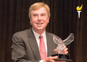 Bill Redpath wearing suit and tie, standing, smiling, holding award resembling a bird, translucent on a small base with a plaque (color photo)